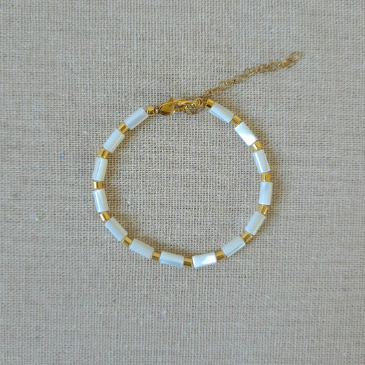 A delicate gold chain bracelet with alternating white rectangular beads and small gold spacer rings, displayed on a textured beige linen background.
